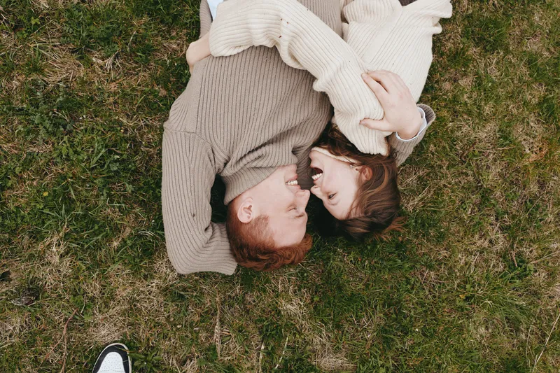 A couple lies on grass, embracing in a cozy outdoor moment, wearing knit sweaters, conveying warmth, connection, and a relaxed lifestyle in a sunlit park setting.
