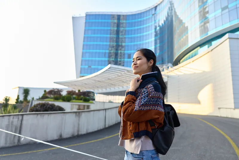 Woman with backpack on an urban pathway beside a modern building, showcasing fashion and travel vibe, outdoor exploration, city architecture under bright daylight and open sky