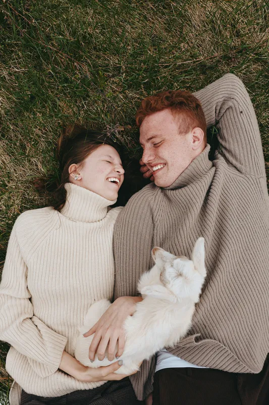 A young couple wearing knitted sweaters lies on the grass smiling while holding a white baby goat, capturing a relaxed and joyful lifestyle moment outdoors in natural light.