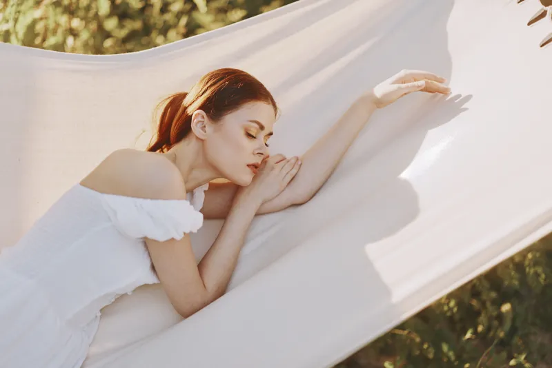 Serene young woman resting on a hammock in a sunny garden, wearing a white dress and reflecting peaceful emotions amidst greenery