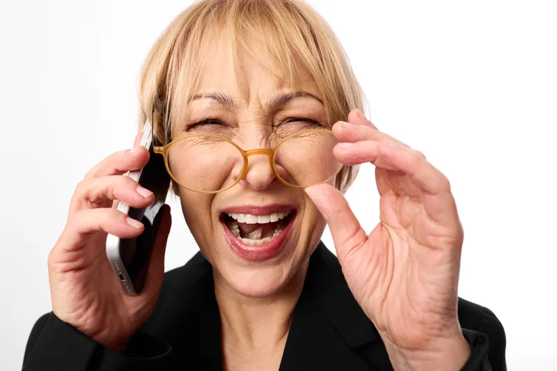 Woman blonde glasses smartphone laughing portrait studio communication business call, holding smartphone by ear, adjusting glasses with hands, bright white background, joyful face