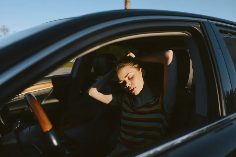Woman lifestyle scene inside a car with soft retro film colors, a relaxed driver rests with hands behind head, striped shirt, sunlit window, moody cinematic vibe