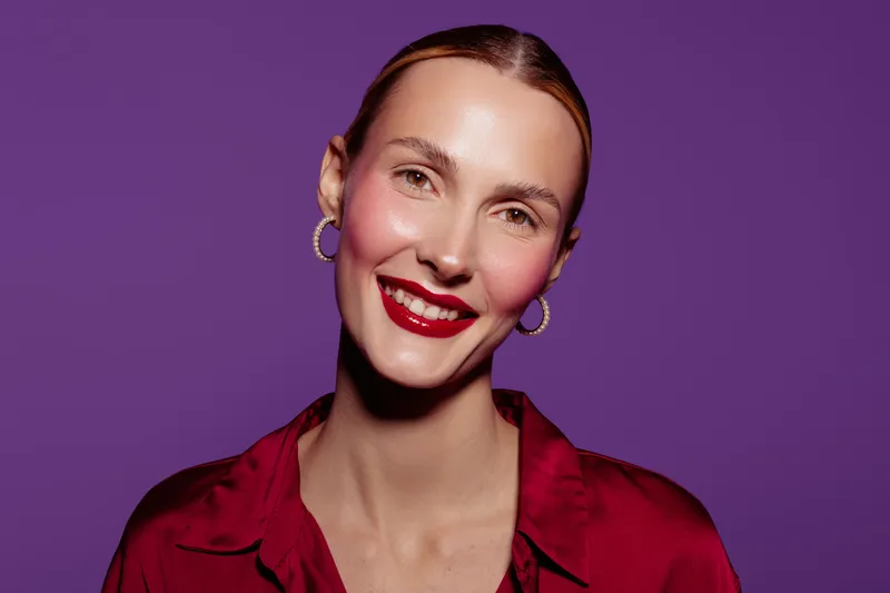 Smiling woman with red lipstick and hoop earrings poses against purple background. She wears a red blouse and has a confident, happy expression. Concept of beauty and fashion.