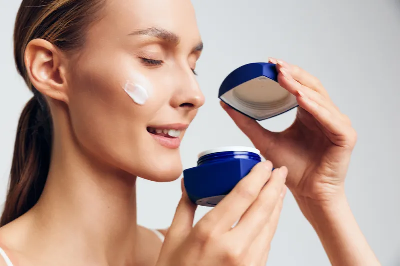 Woman applying face cream moisturizer during skincare routine, holding blue jar and lifting lid, calm close up on healthy skin concept for self care and beauty