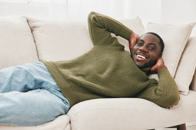 Happy black man relaxing on a sofa in a cozy living room, wearing a green sweater and jeans, conveying comfort and joy in a modern, bright space