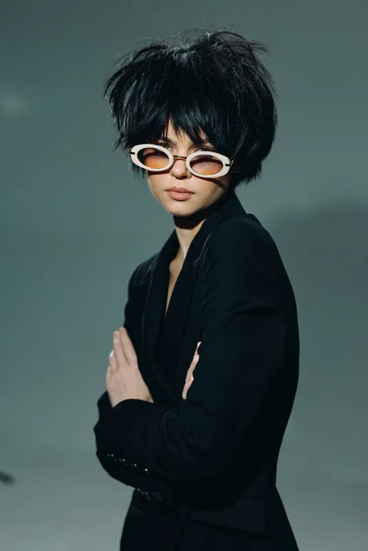 Stylish woman with short black hair wearing white sunglasses and black outfit in moody lighting