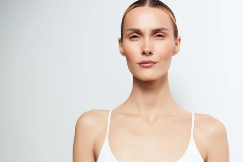 Portrait of confident young woman with natural makeup and blonde hair wearing white tank top looking straight at camera against plain background.
