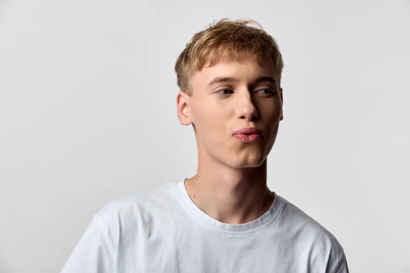 Young man lips expression casual white t shirt in studio light mood close up fashion head shot, candid and simple, neutral background, calm vibe and contemporary styling