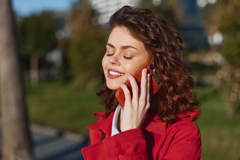 Smiling young woman with curly hair speaks on a red phone in a sunny urban park, conveying friendly everyday communication.