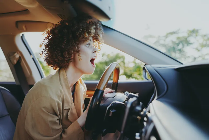 Curly haired woman driving a car with surprised expression, lifestyle moment, warm retro film color, casual outfit, soft lighting, everyday travel scene, candid mood