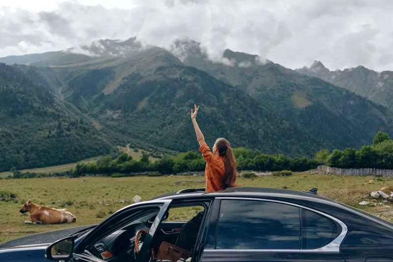 woman car mountains meadow cow roadtrip nature travel sunroof, woman standing through sunroof in meadow with cow and peaks, authentic inclusivity joyful freedom relaxed smile