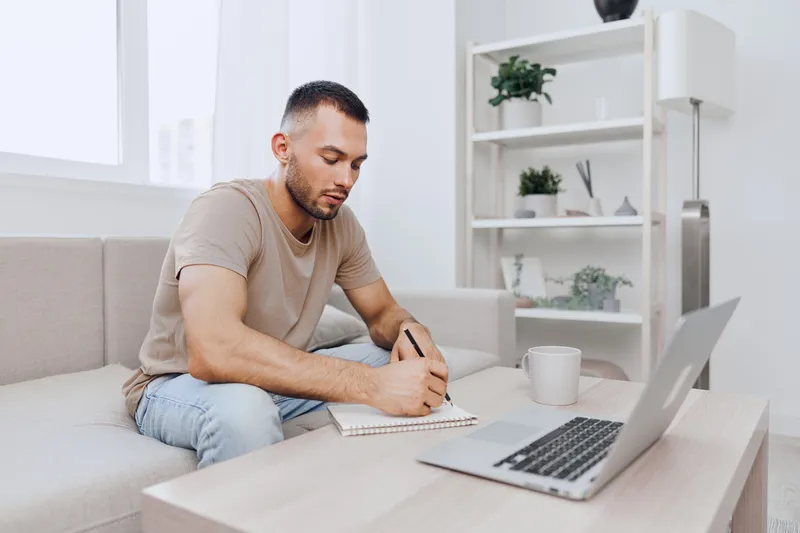 Focused man writing in a notebook at home, surrounded by green plants and natural light. Perfect for themes of productivity and modern living.