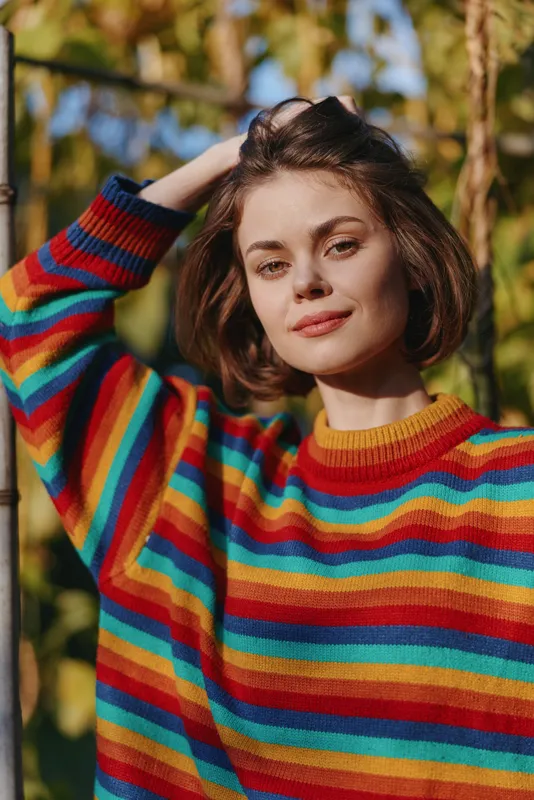 Woman portrait of a young 20s model in a striped sweater outdoors, smiling with short hair and hand on head in an autumn park, casual outfit for lifestyle and fashion use.