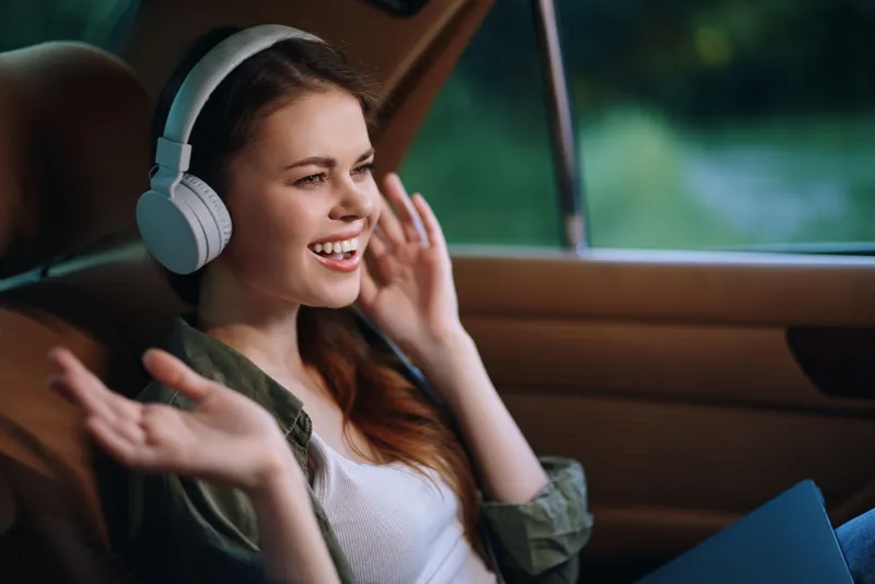 Happy young woman enjoying music with headphones in a car, surrounded by nature, showcasing joy, casual wear, and a relaxed atmosphere.