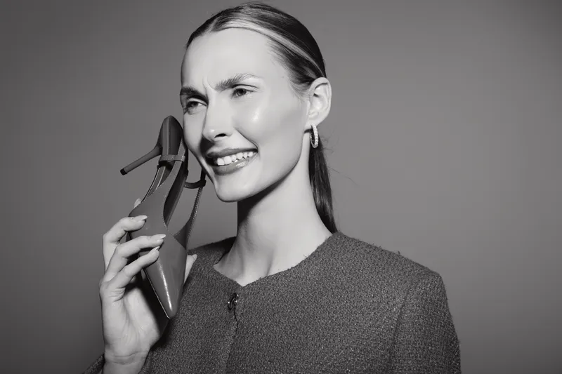 Young white woman with light skin holds high heel shoe near face smiling. Studio portrait on grey background. Concept of fashion, elegance, and modern style.