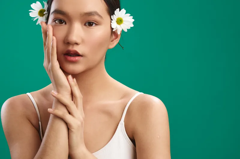 Asian woman with daisies in her hair stands softly against a vibrant green backdrop, exuding gentle elegance and a creative mood. The portrait captures warmth, poise, and a bright, approachable vibe