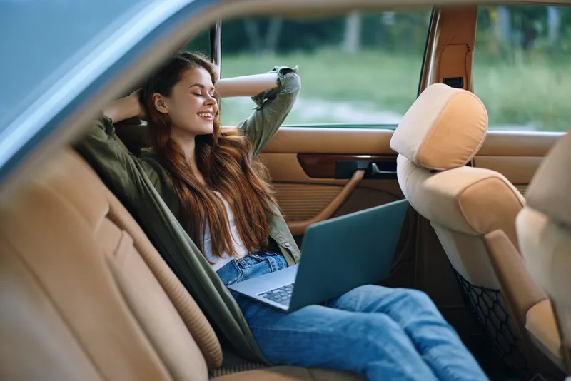 Relaxed young woman enjoying work on her laptop in a stylish car, dressed casually with a joyful expression, surrounded by a natural green backdrop.