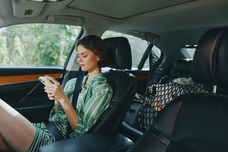 Young woman sits in the car seat, absorbed in her smartphone, wearing a casual striped dress, soft daylight filling the interior.