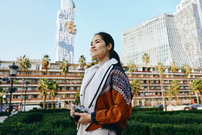 Photographer standing outdoors in an urban setting with camera, scarf and warm jacket, gazing toward the skyline among palm trees and modern glass buildings.