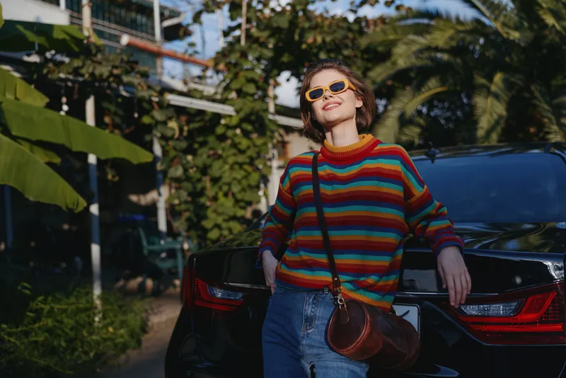 woman in her twenties wearing a colorful striped sweater and sunglasses, leaning on a black car in a tropical street, smiling in a casual outfit with crossbody bag and jeans for travel lifestyle