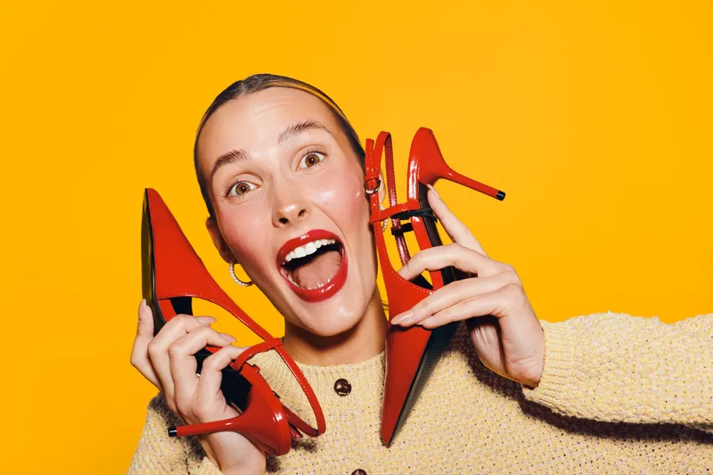 Excited woman with red lipstick holds red high heels near face on bright yellow background. Concept of fashion, style, shopping, happiness, and trendy accessories for women.