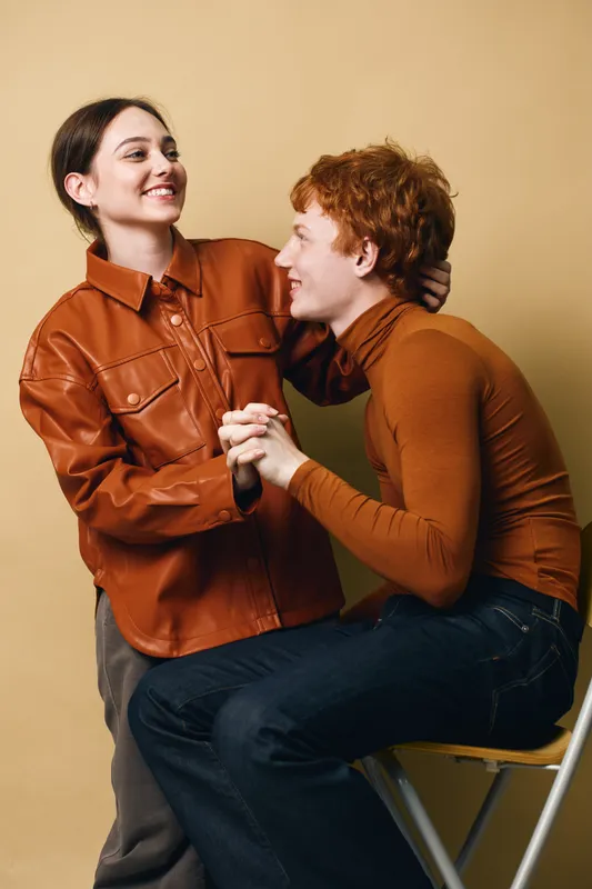 Couple smiling and holding hands with woman standing and man sitting on chair against beige background in casual clothing. Affection, friendship, love, emotions and relationship.