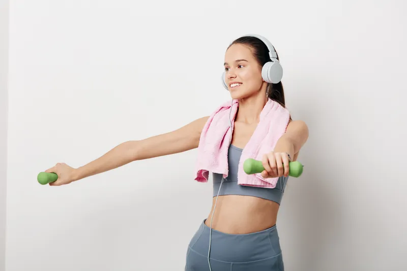 Fitness woman exercises with dumbbells, wearing headphones and a towel draped over shoulders in a clean studio. A dynamic workout moment highlighting strength, balance and focus during a personal