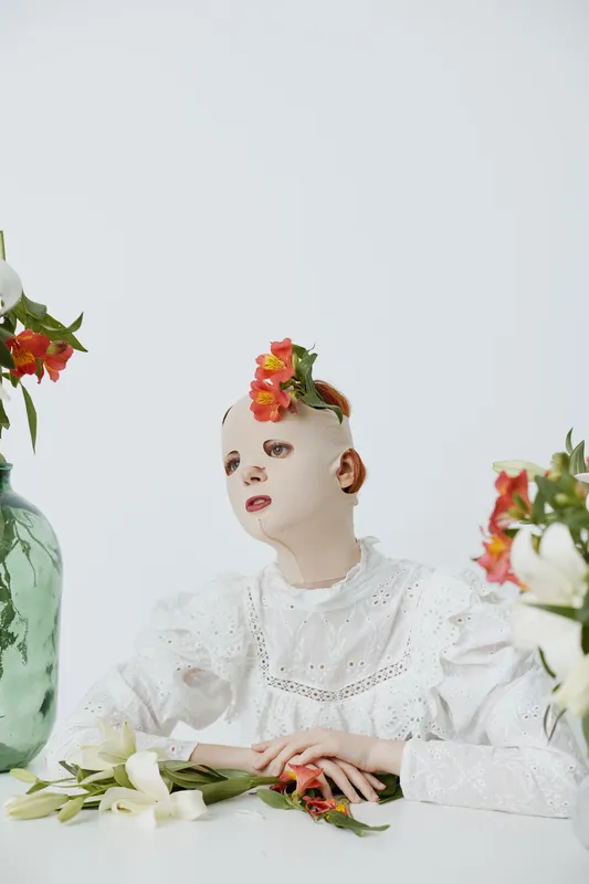 A surreal portrait of a masked model adorned with flowers, sitting in a minimalist setting, evoking emotions of contemplation and beauty