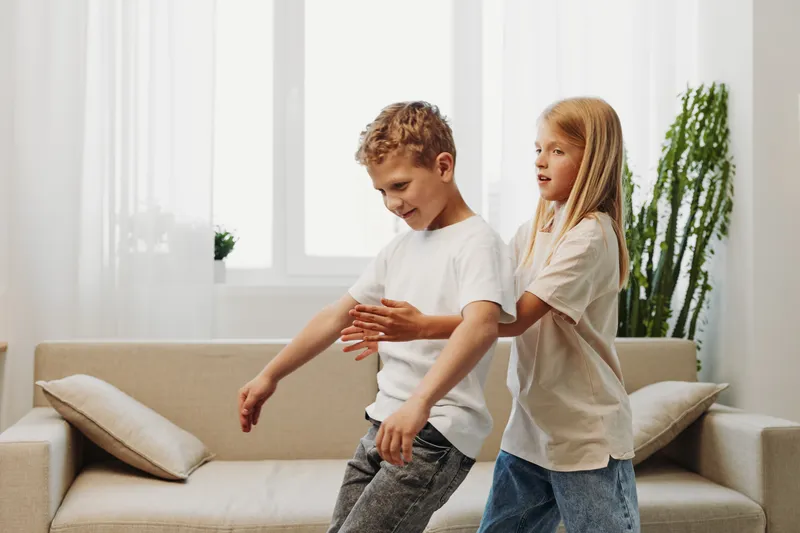 Two children in casual clothes play and laugh in a bright living room with a beige sofa and plants. The boy and girl interact joyfully, creating a warm, playful family atmosphere indoors.