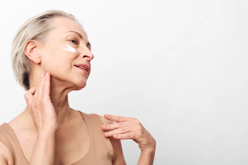 Mature woman with short hair applies face cream on cheek and neck, showing skincare routine and healthy aging on plain white background. Concept of beauty and self-care.