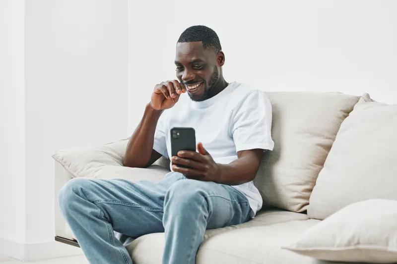 Smiling young Black man enjoying his smartphone while sitting on a light sofa, dressed casually in a white t shirt and jeans in a bright, modern living room