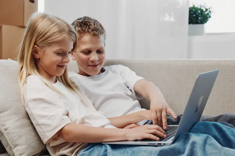 Two happy children sitting on sofa using laptop together in bright room. Concept of technology, friendship, childhood, education, and digital learning at home environment with casual clothes.