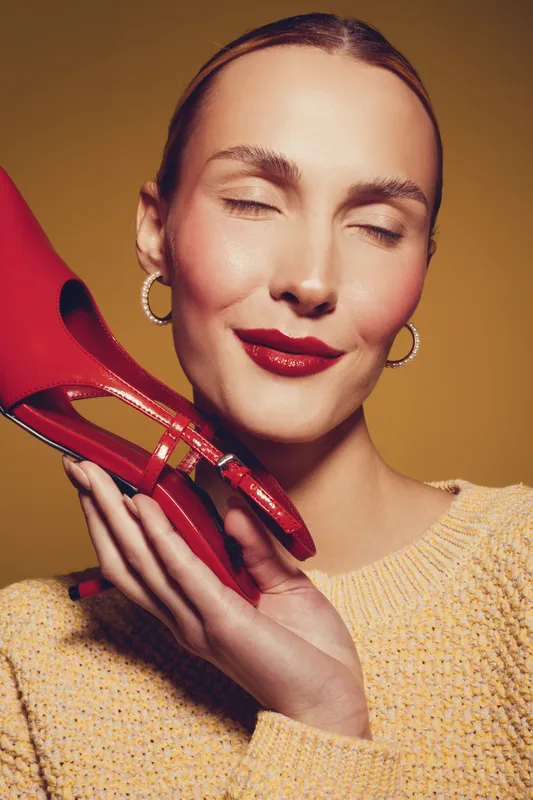 Young woman with closed eyes holds red high heel shoe near face, wearing yellow sweater and hoop earrings. Concept of fashion, style, beauty, and elegance in studio lighting.