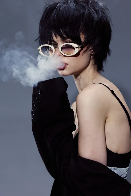Stylish woman with short black hair and sunglasses exhales smoke wearing black jacket and strap dress
