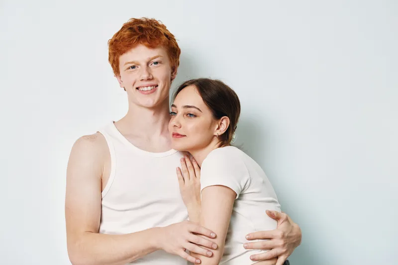 Young couple embracing and smiling together, casual clothing with white shirts on plain background, happy and affectionate portrait of romantic relationship.