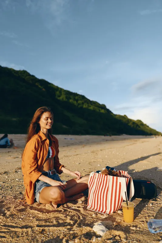 Cheerful woman outdoors on a sunny beach, sitting on sand with a striped bag, picnic setup, orange shirt, drink, and relaxed pose amid green hills and blue sky
