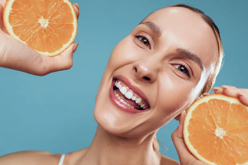 Happy smiling woman holds two halves of fresh orange fruit near face isolated on blue background with natural skin and positive expression in studio shot.