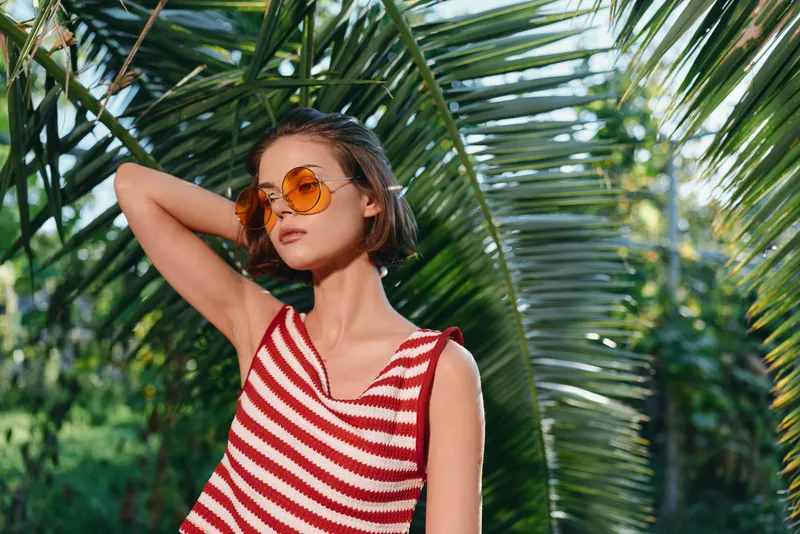 A stylish woman enjoys a sunny day outdoors among lush palm fronds. She wears a red and white striped sleeveless top and orange sunglasses, smiling softly and exuding relaxed confidence in a tropical