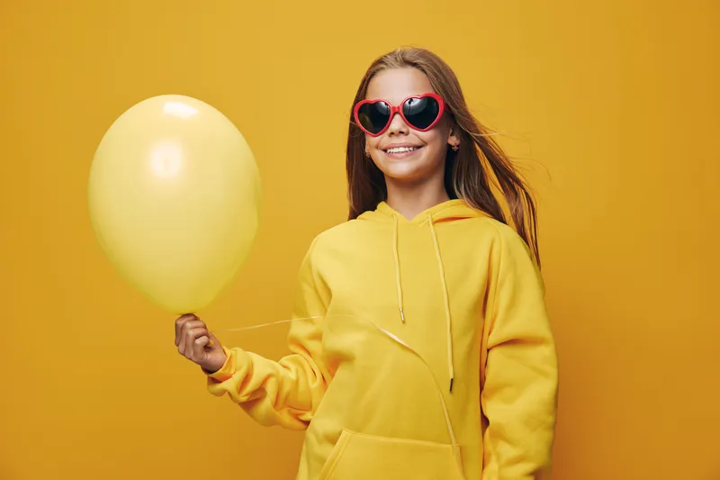 Girl in yellow hoodie holding yellow balloon and wearing red heart shaped sunglasses standing against plain yellow background with smiling pose and casual style.