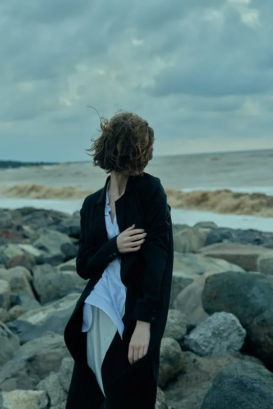 Coastal wind, rocky shore, black coat, white shirt, windy scene, moody atmosphere, solitary figure, stormy sky, ocean, dramatic seascape, fashion mood, windblown details