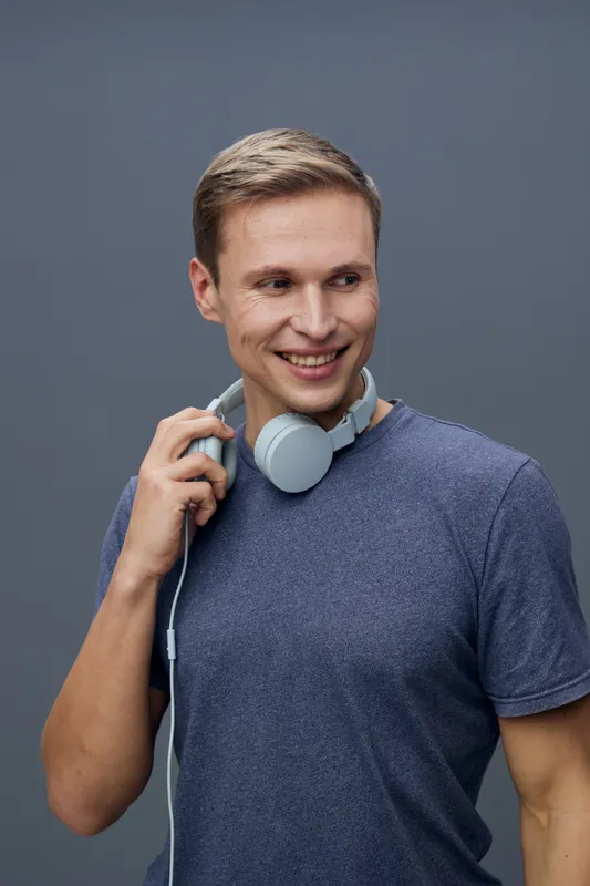 Smiling young caucasian man wearing casual blue t-shirt holding headphones over gray background, looking cheerful and relaxed, studio portrait, lifestyle concept, happiness, music