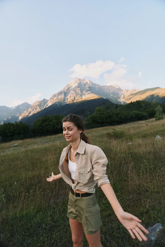 Young woman enjoys open air in a mountain meadow, wearing casual outdoor clothes, arms wide, smiling, capturing carefree lifestyle moments in nature