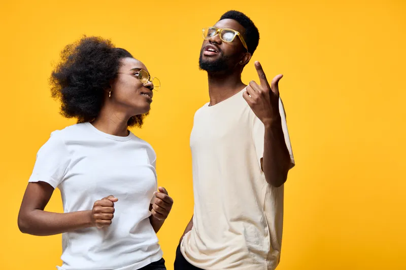 Two joyful young models with afro hairstyles wearing casual outfits and sunglasses, dancing against a vibrant yellow background, expressing happiness and youthfulness