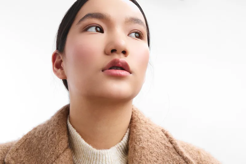 Asian woman gazes upward with a calm expression, wearing a cozy coat, in soft studio light that highlights her thoughtful mood and subtle confidence.