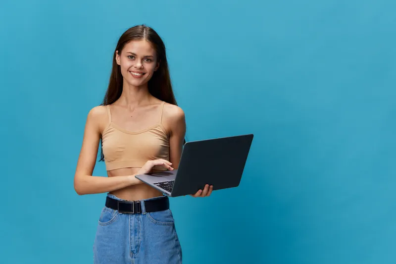 Smiling woman holding laptop stands confidently against blue background with casual clothes, relaxed posture, and natural lighting for a modern tech vibe.