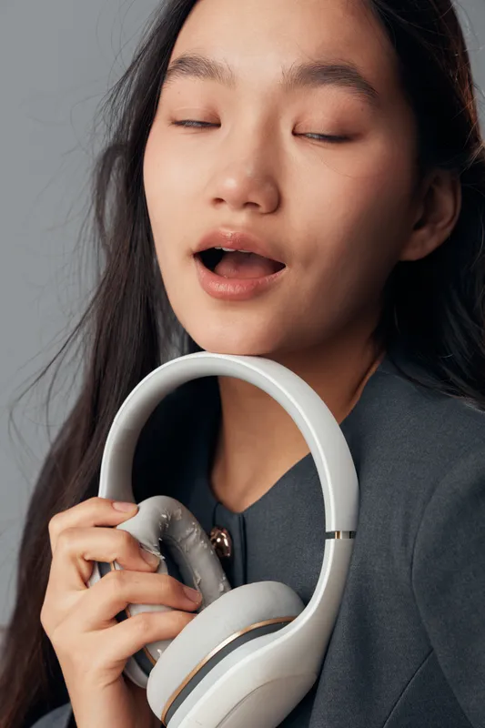 Young woman holding white headphones near face with eyes closed and mouth slightly open in studio. Casual female model with long hair wearing dark clothing against gray background.