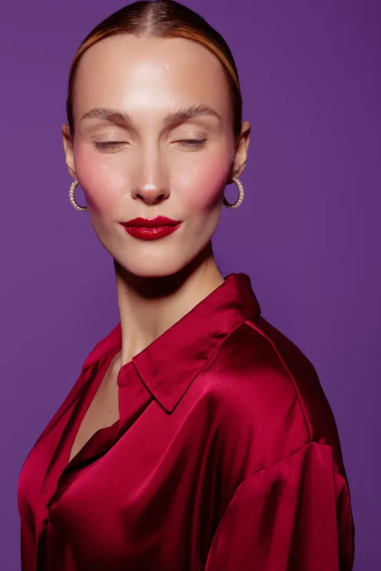 Close-up portrait of a woman with closed eyes wearing red lipstick and satin blouse against purple background. Concept of beauty, fashion, elegance, and makeup artistry.