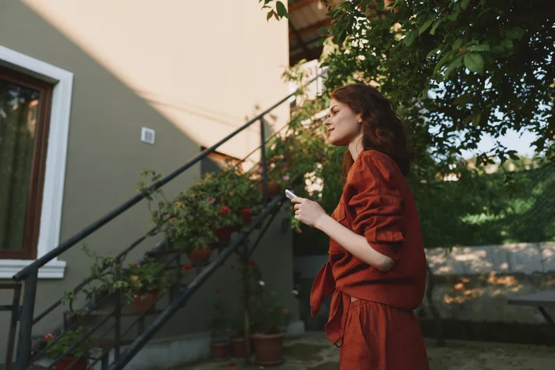 Woman with curly hair in a red outfit stands by outdoor steps, looking up. Natural light creates a warm, relaxed lifestyle moment with greenery and urban charm