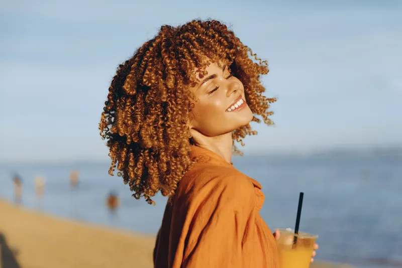 Smiling woman with curly hair holding a cold drink on a sunny beach, with blue water behind. Casual lifestyle, wellness, and summer vibes presented in bright, natural light.