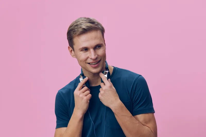 Smiling man with headphones, casual blue t-shirt, happy expression, young adult, isolated on pink background, studio portrait, cheerful mood, relaxed style, lifestyle concept, positive energy.